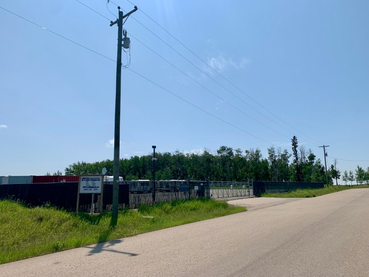 Entrance to Trailing West RV and Boat Storage facility between Sundre and Olds, Alberta, showing secure gated access, chain-link fence, security cameras, and spacious gravel lot for RV and trailer parking.